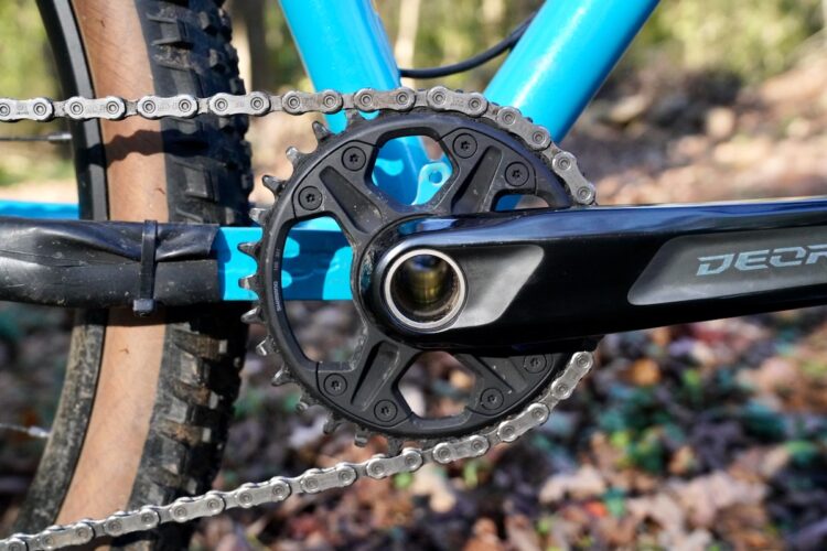 Close-up view of a bicycle's drivetrain, featuring a black crankset with a round chainring and a silver chain. The background includes a blurred forest floor with leaves, and the bike frame is shown in blue.