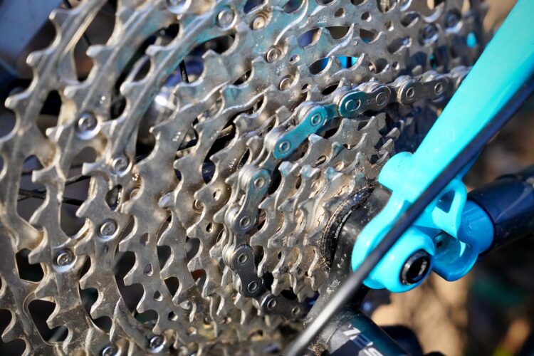 Close-up of a bicycle cassette and chain, featuring gleaming metallic gears and a turquoise frame component, with a focus on the intricate details of the chain links and sprocket design.