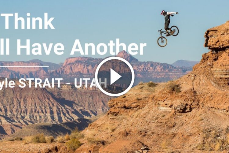 A mountain biker performing a jump off a rocky cliff in a desert landscape, with colorful mountains in the background and text overlay that reads "I Think I'll Have Another" and "Kyle STRAIT - UTAH." A play button indicates the presence of a video.