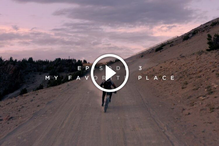 A cyclist rides along a dirt road at dusk, surrounded by a landscape of rocky terrain and sparse vegetation. The sky features soft hues of pink and purple. Text overlay reads "EPISODE 3 MY FAVORITE PLACE" with a play button in the center.