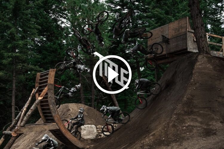 A montage of a mountain biker performing multiple aerial tricks over a dirt ramp, surrounded by tall trees. The biker is captured in various stages of the jump, showcasing dynamic movement and skill. A play button graphic is overlayed on the image, suggesting it is part of a video.