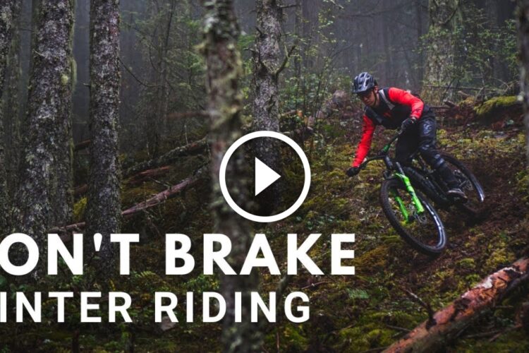 A mountain biker in a red long-sleeve shirt and black gear navigates a curved trail through a misty forest, surrounded by tall trees and moss-covered ground, with the text "DON'T BRAKE WINTER RIDING" displayed prominently. A play button icon is included, indicating the image is a thumbnail for a video.
