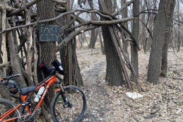 A mountain bike resting against a tree near a whimsical wooden structure with a sign that reads "FART CITY." The surrounding area features bare trees and a leaf-covered ground, indicating a late autumn or early spring setting. A dirt path leads away from the structure into the wooded landscape. ESU Trail mountain bike trail.