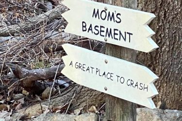 A humorous wooden sign with two directional arrows: the top arrow reads "MOM'S BASEMENT" and the bottom arrow states "A GREAT PLACE TO CRASH," surrounded by natural foliage and tree branches. Indigo Loop mountain bike trail.