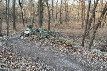 A winding dirt path in a wooded area, surrounded by bare trees and autumn leaves. To the side of the path, there is a stone structure made of stacked rocks. The scene is set in a natural landscape, indicating a tranquil outdoor environment. Indigo Loop mountain bike trail.
