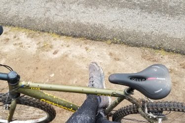 Image of a person sitting on a bicycle with muddy legs, wearing black leggings and gray shoes. The bike is green with yellow detailing and is positioned next to a gravel path. The surroundings show a grassy area and an unpaved surface. Mercer County Park mountain bike trail.
