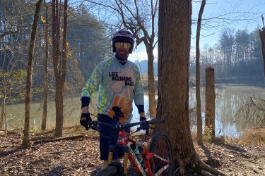 A mountain biker wearing a colorful jersey and helmet stands next to a red bicycle in a wooded area near a lake. The background features bare trees under a clear blue sky, and the ground is covered with leaves. Warrior Creek mountain bike trail.