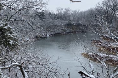 A tranquil river scene surrounded by snow-covered trees, with branches dusted in white against a gray sky. The calm water reflects the wintry landscape, creating a serene winter atmosphere in a natural setting. Lehigh Portland Trails mountain bike trail.
