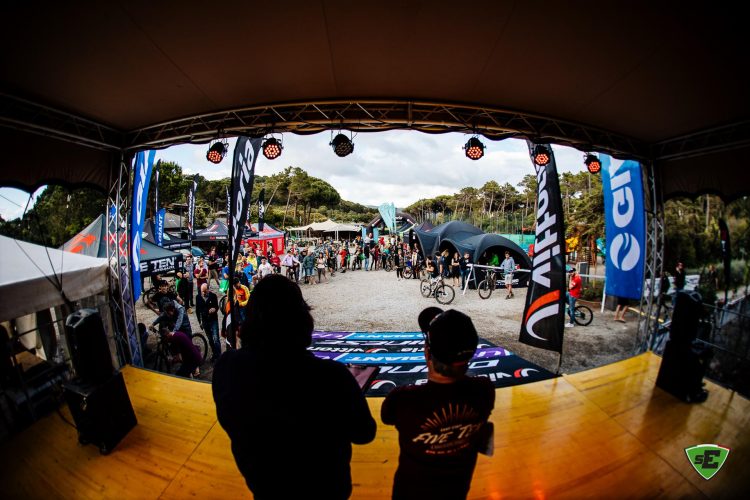 A view from a stage overlooking a large crowd at an outdoor event. Attendees are gathered around various vendors' tents displaying biking equipment and merchandise. Flags representing different brands are prominently displayed, and some individuals are engaging with mountain bikes. The scene is lively, with trees in the background and a cloudy sky above.