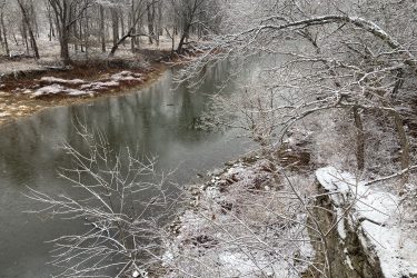 A serene winter landscape showcasing a calm river bordered by snow-covered trees. The branches are frosted with ice, and a light dusting of snow blankets the ground, creating a peaceful and quiet atmosphere. The water reflects the surrounding scenery, contributing to the tranquil scene. Lehigh Portland Trails mountain bike trail.