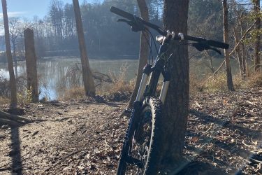 A mountain bike leaning against a tree by a peaceful lake, surrounded by bare trees and fallen leaves, with sunlight shining through. The scene captures a serene outdoor environment ideal for biking and nature experiences. Warrior Creek mountain bike trail.