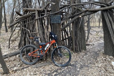 A mountain bike is parked next to a rustic wooden structure made of branches, marked with a sign that reads "FART CITY." The surrounding area is covered in fallen leaves, and several bare trees are visible in the background, suggesting an outdoor wooded environment. ESU Trail mountain bike trail.