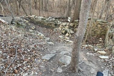 A narrow dirt path winding through a forest, bordered by autumn leaves and bare trees. In the background, a weathered stone wall with some loose stones is visible, indicating the remnants of an old structure. The scene is quiet and natural, evoking a sense of tranquility in a wooded area. Indigo Loop mountain bike trail.