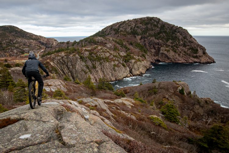 A mountain biker navigates rocky terrain overlooking a coastal landscape with cliffs, trees, and the ocean in the background, under a cloudy sky.