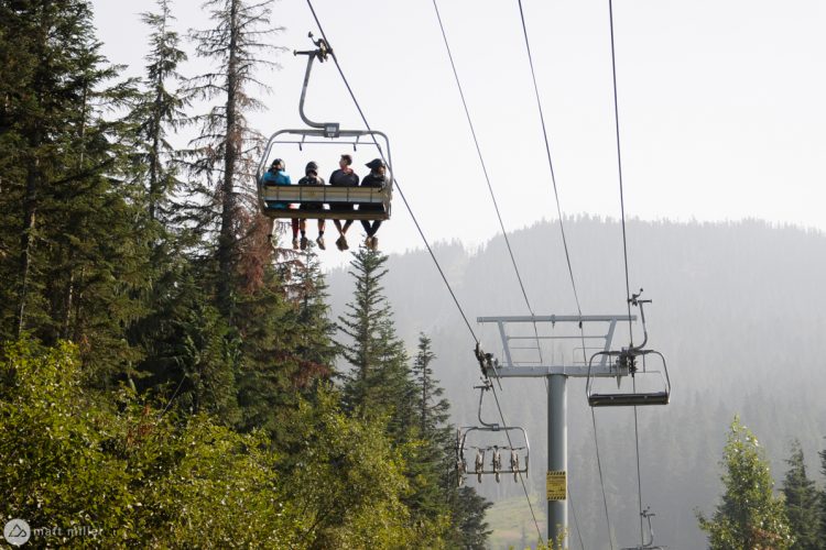 Four people riding a ski lift above a dense forest, surrounded by tall evergreen trees and misty mountains in the background. The lift cables are visible, and the scene captures a peaceful outdoor atmosphere.