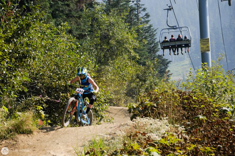 A mountain biker navigates a dirt trail surrounded by greenery, with a ski lift carrying passengers in the background. The biker is wearing a helmet and racing gear, and the trail features a slight incline, suggesting a dynamic biking environment.