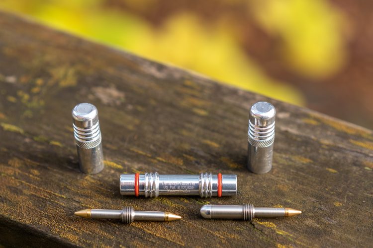 A collection of metallic pen components arranged on a wooden surface, featuring two outer pieces, a central cylindrical piece with an orange ring, and two gold-tipped pen nibs. The background is softly blurred with hints of yellow, suggesting an outdoor setting.