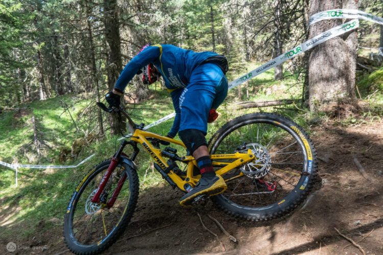 A mountain biker in a blue and yellow outfit leans into a turn on a trail surrounded by trees, demonstrating a dynamic riding position on a yellow bike with red accents. A race banner is visible in the background. The scene captures the excitement of mountain biking in a forested setting.