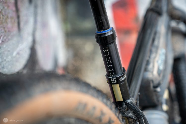 Close-up image of a mountain bike's dropper post, showing the measurement markings and adjustment mechanism. The background features blurred graffiti, adding a colorful urban context to the scene. The bike exhibits signs of use with dirt on the tire, emphasizing an adventurous outdoor setting.