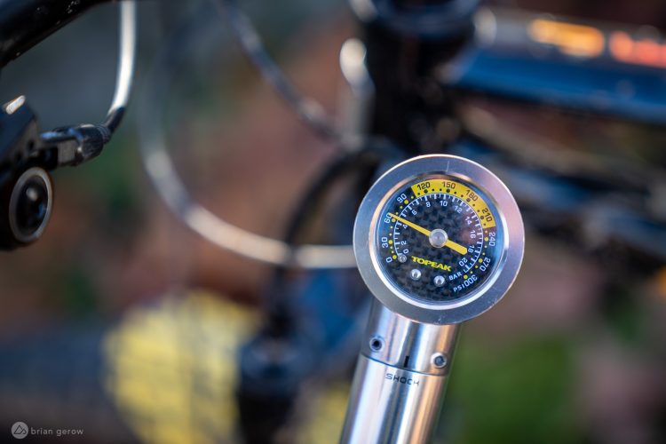 A close-up of a Topeak pressure gauge attached to a bicycle, displaying pressure measurements in psi and bar, with a blurred background featuring bicycle components and natural elements.