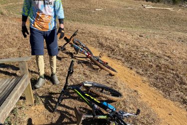 A mountain biker wearing a colorful jersey and helmet stands on a dirt path with two bicycles resting nearby, one partially on its side. The background features a bare field and trees, indicating a natural outdoor setting. Warrior Creek mountain bike trail.
