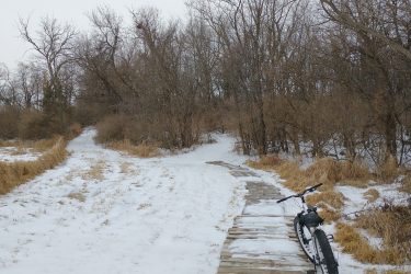 A winter scene featuring a snow-covered path leading into a wooded area. On the right side, a wooden boardwalk is visible, and a bicycle rests on the ground next to it. Bare trees can be seen in the background under a cloudy sky, with patches of snow and dried grass surrounding the path. Lake LaShane Trail mountain bike trail.
