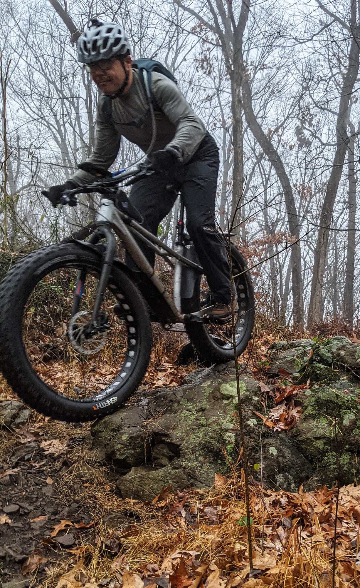 A person riding a mountain bike is navigating a rocky trail in a misty forest. The rider is focused as they maneuver over a large boulder, with autumn leaves scattered on the ground and trees in the background. Watchung mountain bike trail.