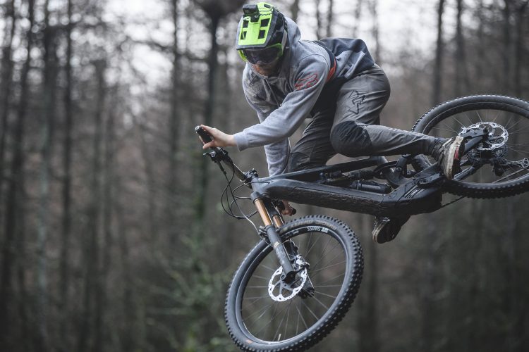 A mountain biker performing a jump in a wooded area, wearing a grey hoodie and green helmet with a camera attached. The biker is airborne, showcasing a dynamic pose on a black mountain bike, surrounded by trees. The scene captures the thrill of extreme sports in an overcast atmosphere.