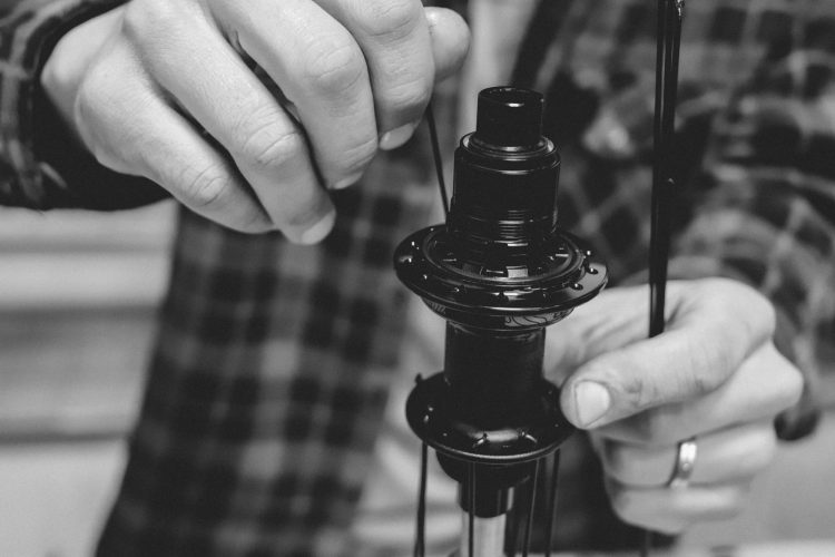 A close-up image of hands working on assembling a black bicycle hub component, with a focus on precise mechanical adjustments and tools present. The background is blurred, emphasizing the intricate details of the hub and the person's hands.