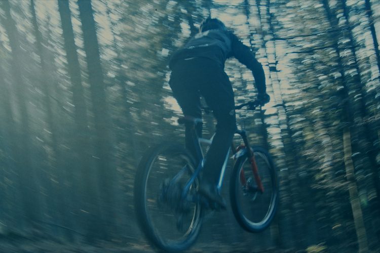 A person riding a mountain bike, captured in motion while jumping through a forest. The image features blurred trees in the background, conveying speed and adventure.