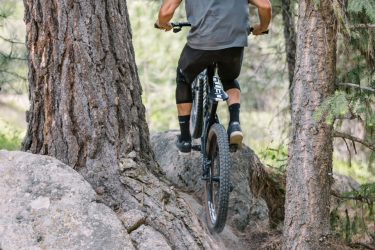 A cyclist riding a mountain bike on a rocky trail surrounded by tall trees, captured from behind. The rider is wearing a helmet and a gray t-shirt, navigating a steep section of the path with visible dust being kicked up.