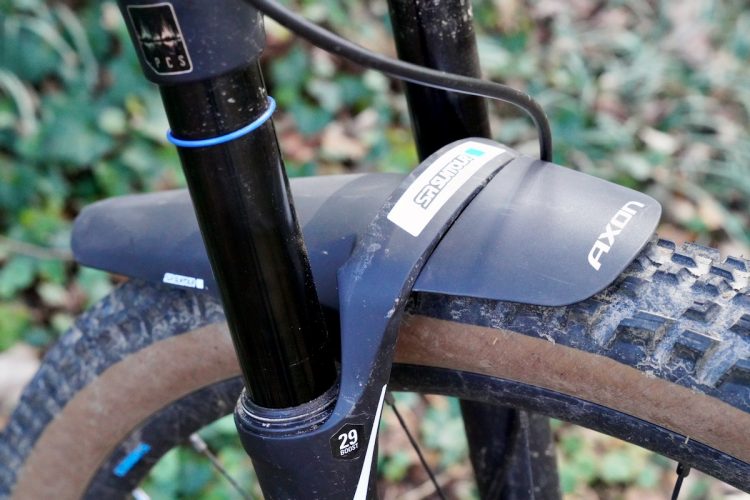 Close-up image of a mountain bike's front fork and tire, featuring a black suspension fork and a mudguard labeled "AXON." The bike has dirty tires and is set against a blurred background of green foliage. A blue rubber ring is visible around the fork, indicating a bike maintenance detail.