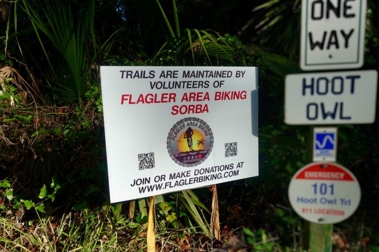 Sign detailing that the trails are maintained by volunteers from Flagler Area Biking SORBA, encouraging donations and participation. The sign includes a logo and QR code, with nearby road signs indicating a one-way direction and the name "Hoot Owl." Lush greenery surrounds the sign.