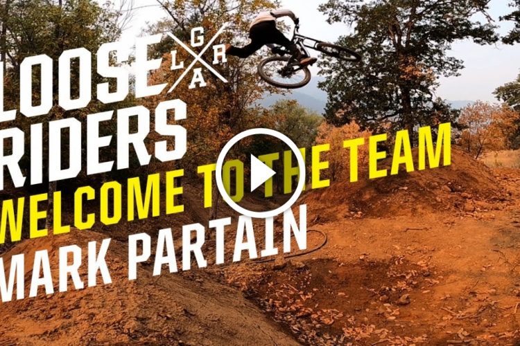 A mountain biker performing a jump over a dirt ramp in an outdoor setting, surrounded by trees and autumn foliage. The image features bold text that reads "LOOSE RIDERS" at the top, along with "WELCOME TO THE TEAM" and "MARK PARTAIN" in bright yellow letters. A play button icon is also visible, indicating that this image is part of a video.