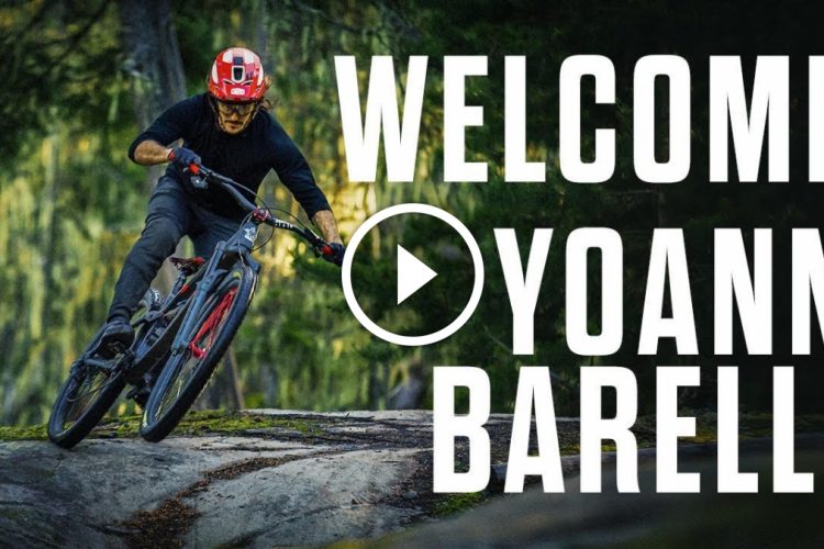 A mountain biker in a black shirt and red helmet is skillfully riding down a rocky trail surrounded by lush green trees. The text "WELCOME YOANN BARELLI" is prominently displayed alongside a play button graphic.