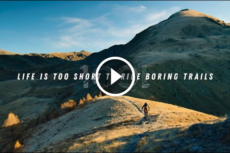 A scenic landscape featuring rolling hills and mountains under a clear blue sky, with a person riding a mountain bike along a winding trail. The image includes the text "LIFE IS TOO SHORT TO RIDE BORING TRAILS" prominently displayed.