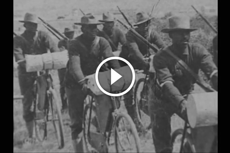 A historical black and white image depicting a group of soldiers riding bicycles, each carrying a wooden crate and armed with rifles. The scene captures a moment from the past, highlighting the soldiers in their military attire as they traverse a rural landscape.