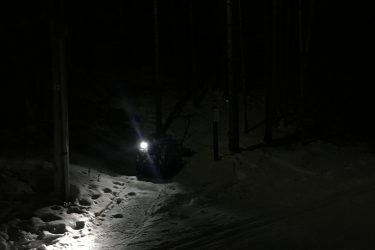 A snow-covered path illuminated by a bright light in a dark forest at night, with tree silhouettes in the background and a snowmobile visible on the side. Pine Valley mountain bike trail.