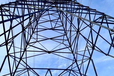 A low-angle view of a tall power transmission tower against a clear blue sky, showcasing the intricate metal framework and overhead power lines extending in various directions. Batawa Trails mountain bike trail.