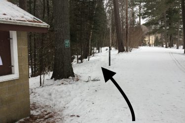 Entrance to a mountain bike trail in a snow-covered forest. The image shows a snow-dusted path alongside trees, with a sign indicating the trail's entrance. A building is partially visible on the left. Pine Valley mountain bike trail.