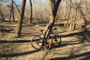A blue mountain bike leaning against a tree on a dirt path in a wooded area. Bare trees line the path, and an orange backpack is resting on the ground near the bike. The scene is illuminated by sunlight, creating a serene outdoor setting. Pryor Creek Nature Trail mountain bike trail.