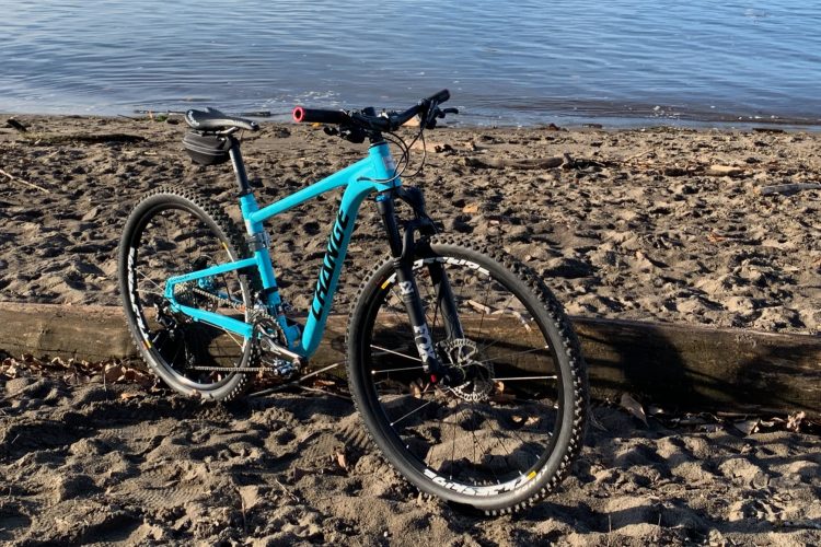 A blue mountain bike is parked on a sandy beach next to a calm body of water, with a log lying in the background. Sunlight illuminates the scene, highlighting the bike's frame and tire details.