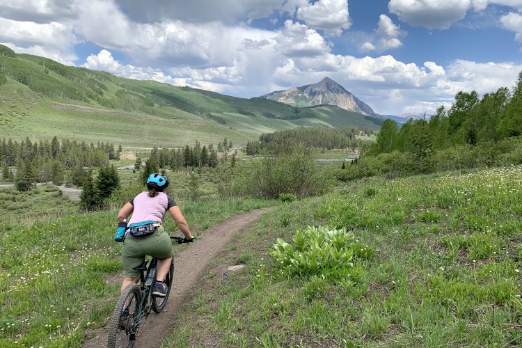 A person riding a mountain bike along a dirt trail in a lush green landscape, with rolling hills and a prominent mountain in the background under a partly cloudy sky. The cyclist is wearing a helmet and athletic clothing, surrounded by trees and wildflowers.