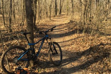 A mountain bike resting against a tree on a narrow trail through a wooded area, with fallen leaves covering the ground and leafless trees in the background. An orange backpack is placed on the ground near the bike. Pryor Creek Nature Trail mountain bike trail.