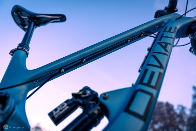 Close-up view of a mountain bike frame with a sleek design, featuring a blue color scheme and the word "DEVIATE" prominently displayed. The image captures details of the seat post and frame geometry against a soft, twilight sky.