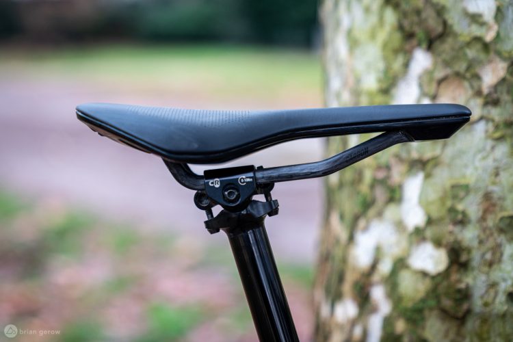 Close-up of a black bicycle saddle attached to a sleek black seat post, positioned next to a textured tree trunk in a blurred outdoor setting. The saddle features a streamlined design with a textured surface, suggesting a focus on comfort and performance.