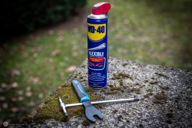 A can of WD-40 with a flexible straw, positioned next to a multi-tool with a blue handle and a socket wrench, resting on a moss-covered stone surface. The background features a blurred, green outdoor setting.