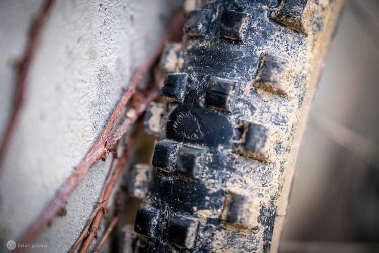 Close-up view of a textured black tire with a knobby tread pattern, partially covered in dirt, against a light gray concrete background with some brown vines intertwined.