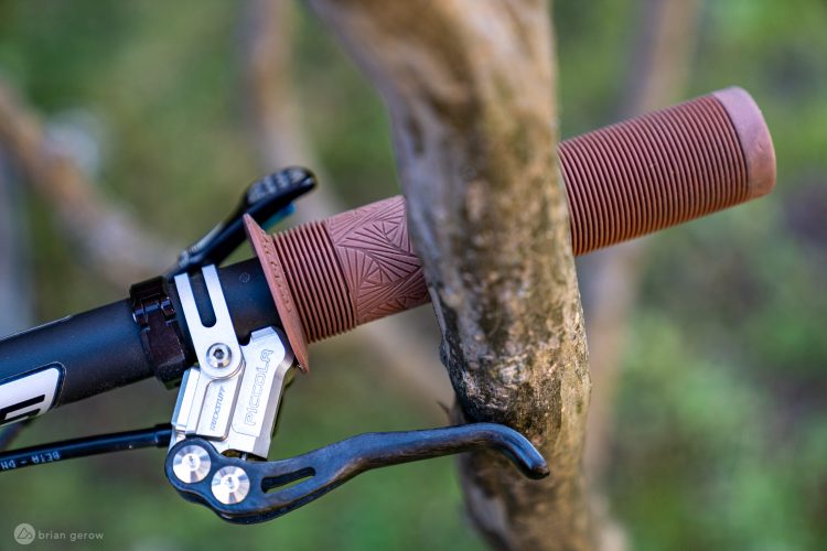 Close-up of a bicycle handlebar grip resting against a tree, featuring a textured brown grip and a silver brake lever with visible components. The background is softly blurred with green foliage, suggesting an outdoor setting.