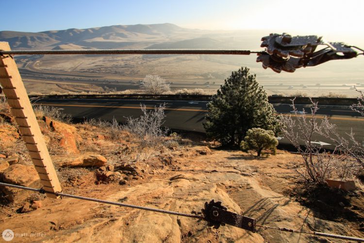 A scenic view from a rocky overlook, featuring a wire fence with a latch, overlooking a winding road below. In the distance, rolling hills are visible, with a light frost covering the vegetation. The sky is clear, suggesting a bright morning atmosphere.